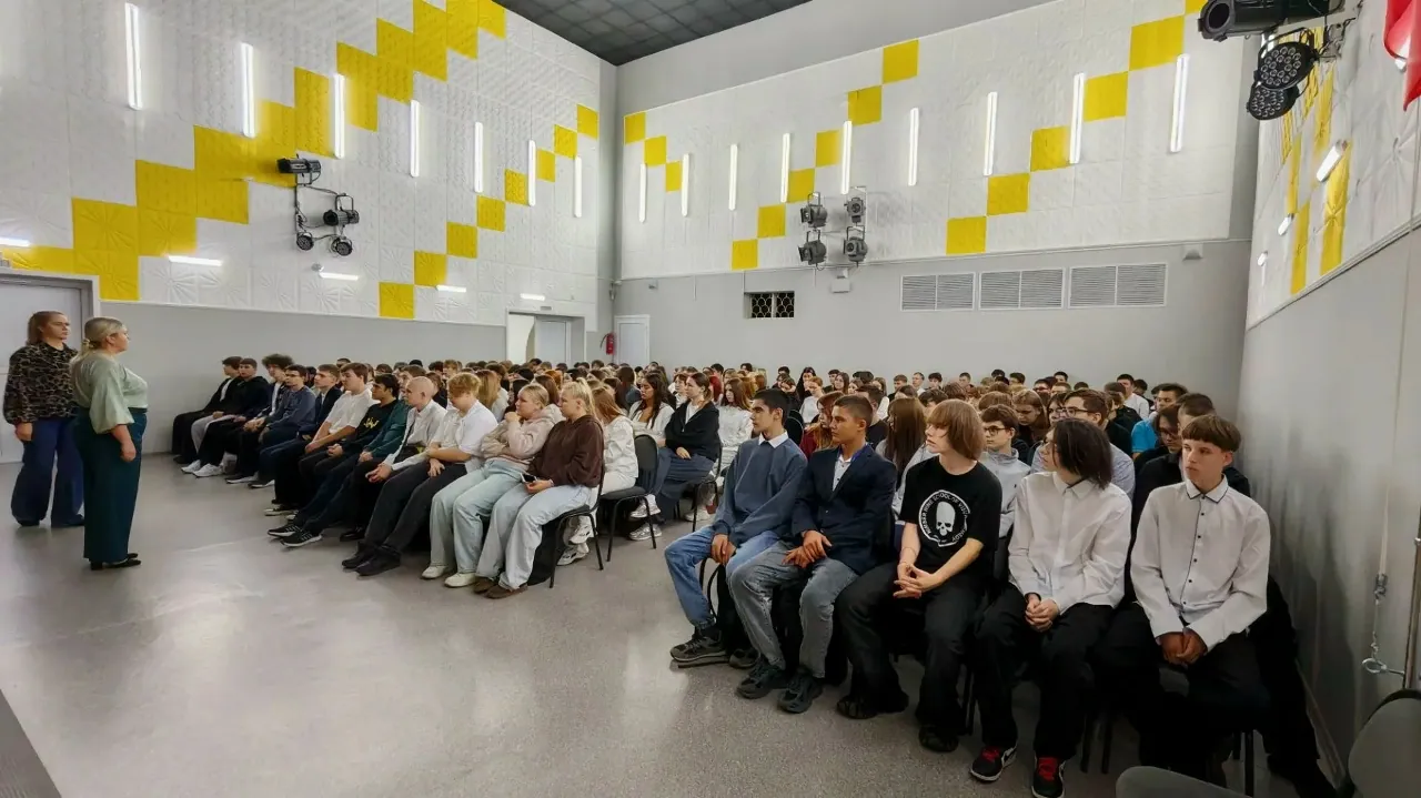Просторное помещение с высокими белыми стенами, украшенными желтым геометрическим орнаментом в виде зигзагов. Слева стоят две женщины (вероятно, преподаватели или организаторы), обращенные лицом к залу. Справа на стульях сидит большая группа молодых людей, студентов или школьников, которые внимательно слушают выступающих. На стенах закреплены светильники и проекционное оборудование, на полу — светлое гладкое покрытие.