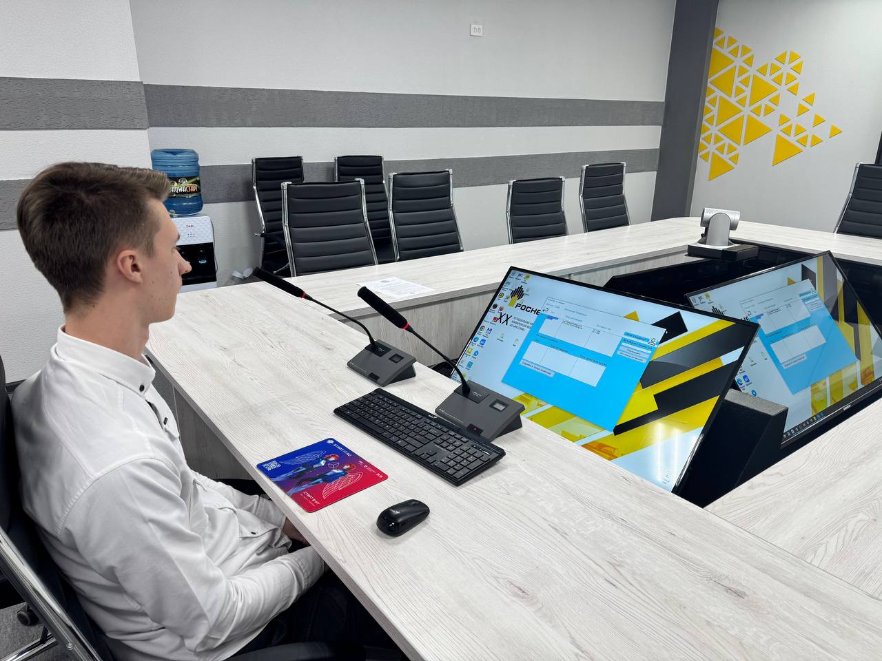 изображен молодой человек, сидящий за столом в конференц-зале. Он одет в белую рубашку и смотрит на большой экран, который находится перед ним. На столе размещены клавиатура и мышь, а также папка с документами. Стена за ним оформлена в светлых тонах с желтыми геометрическими узорами. В помещении много серых стульев, расположенных вокруг стола.