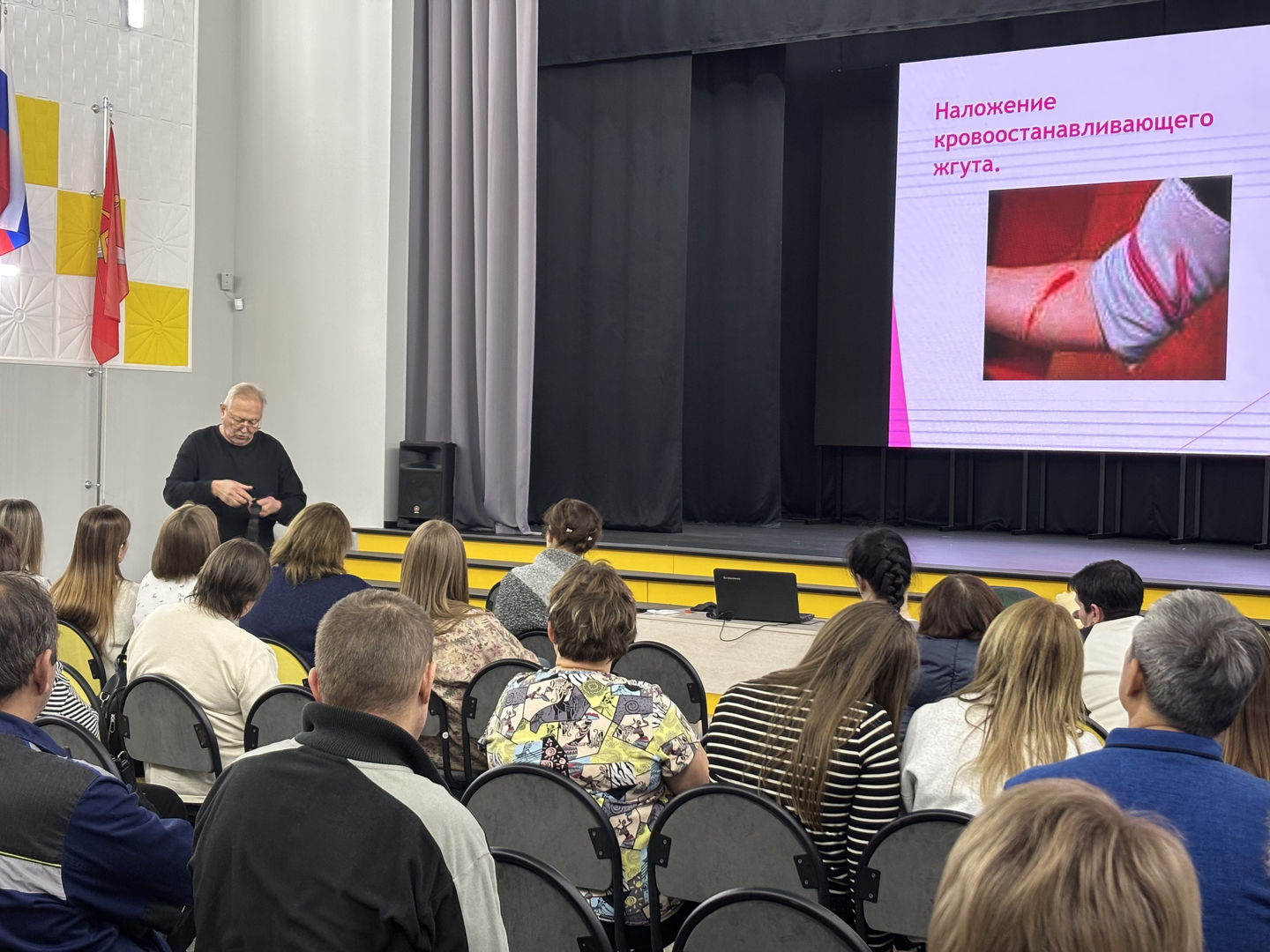 видно мужчину, который стоит и демонстрирует наложение кровоостанавливающего жгута на своем предплечье. Снимок сделан с небольшой дистанции, чтобы хорошо видно было как жгут применяется. На заднем плане — экран с презентацией, где видно изображение жгута и текст, связанный с темой. Аудитория состоит из людей, внимательно слушающих и смотрящих на спикера, некоторые из них делают заметки.