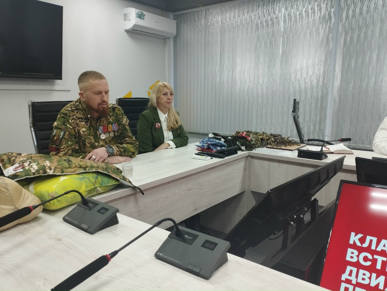 На этом фото два человека находятся в офисе. Слева сидит Борис Филиппов в камуфляжной форме с медалями на груди и серьёзным выражением лица. Справа от него женщина с длинными светлыми волосами, одетая в зелёный пиджак. Оба смотрят в сторону стола, на котором разбросаны различные тканевые материалы и устройства. В офисе присутствует современное оборудование, а на заднем плане виден телевизор и окна с прозрачными шторами.