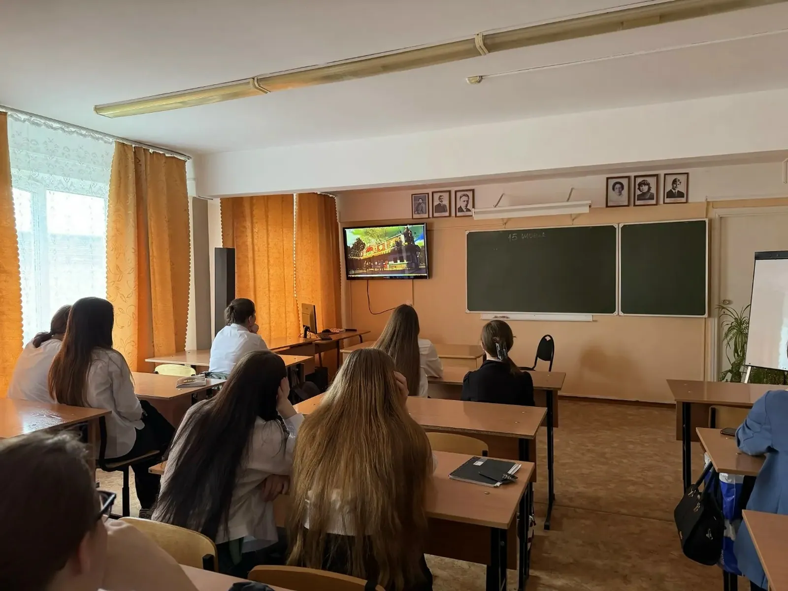 Ученики в классе, сидящие за рядами парт и смотрящие на телевизор у доски; в помещении шторы, плакаты и школьная мебель.