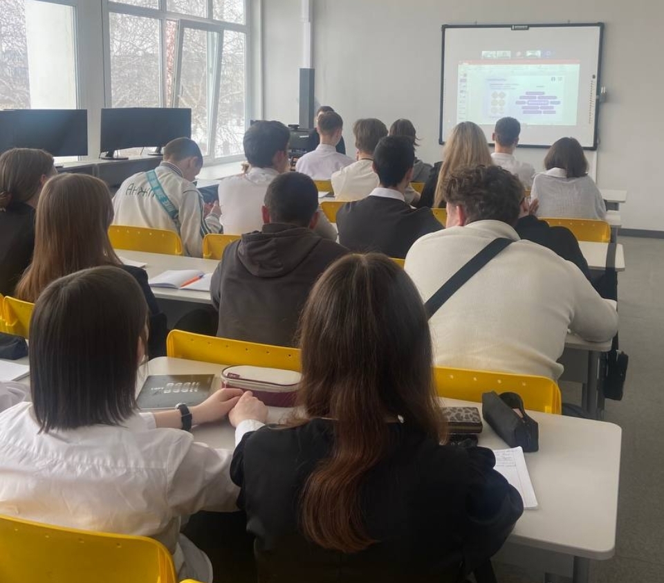 запечатлен класс, однако фокус сменился на заднюю часть аудитории. Студенты, сидящие за столами, обращены к экрану, на котором отображается какая-то презентация. Большинство учащихся одеты в светлую одежду, многие из них поддерживают друг друга, держа за руки. Атмосфера сосредоточенности и внимательности, с вниманием обращенным на то, что происходит на экране.