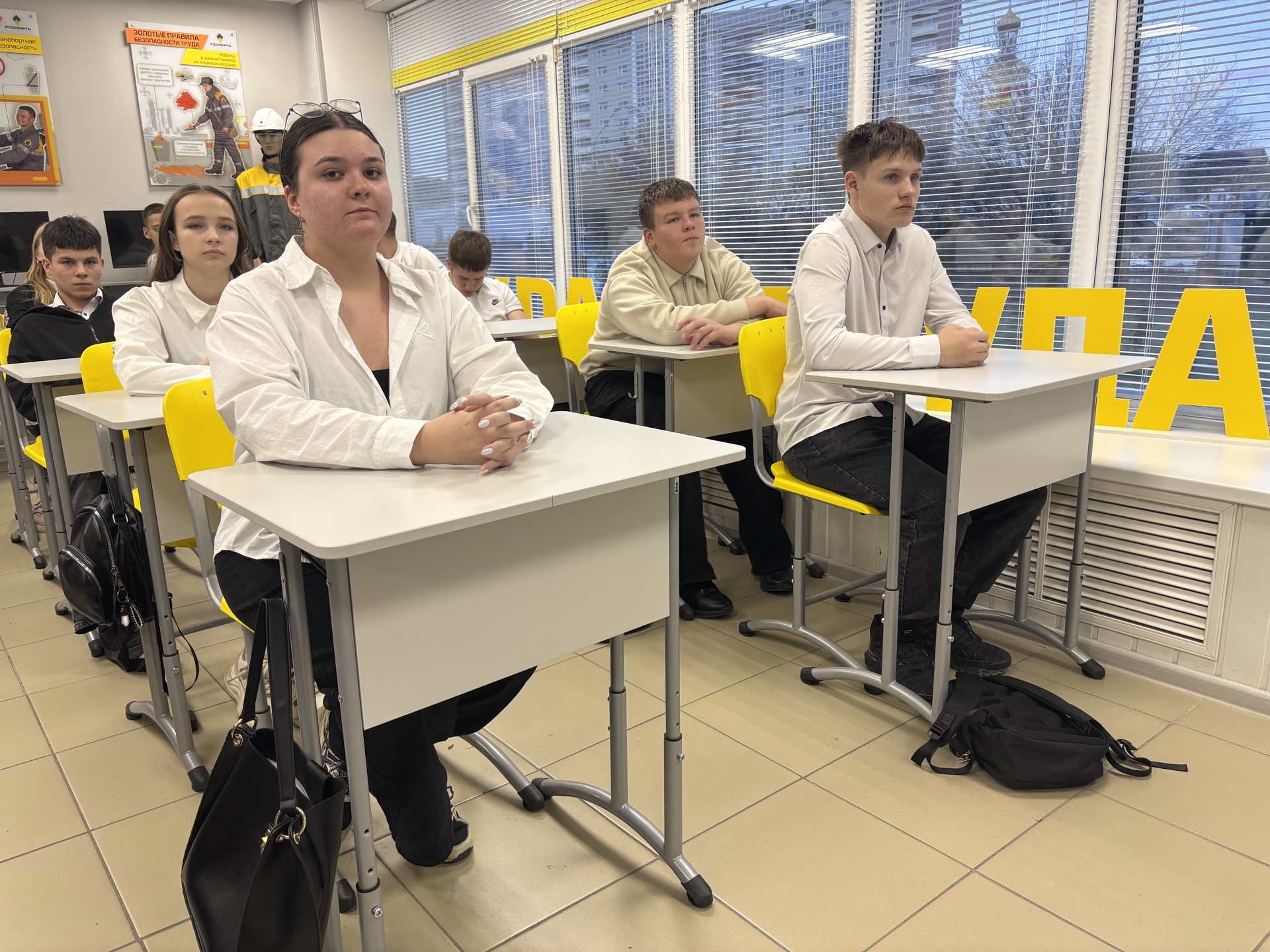 На изображении представлена группа учащихся, сидящих за столами в классе. Все они одеты в белые рубашки, а фон классной комнаты включает желтые стулья и окна с жалюзи. Учащиеся внимательно смотрят вперед, сосредоточенные на чем-то, что происходит в классе. Комната имеет яркое освещение, а на стенах видны некоторые учебные материалы.