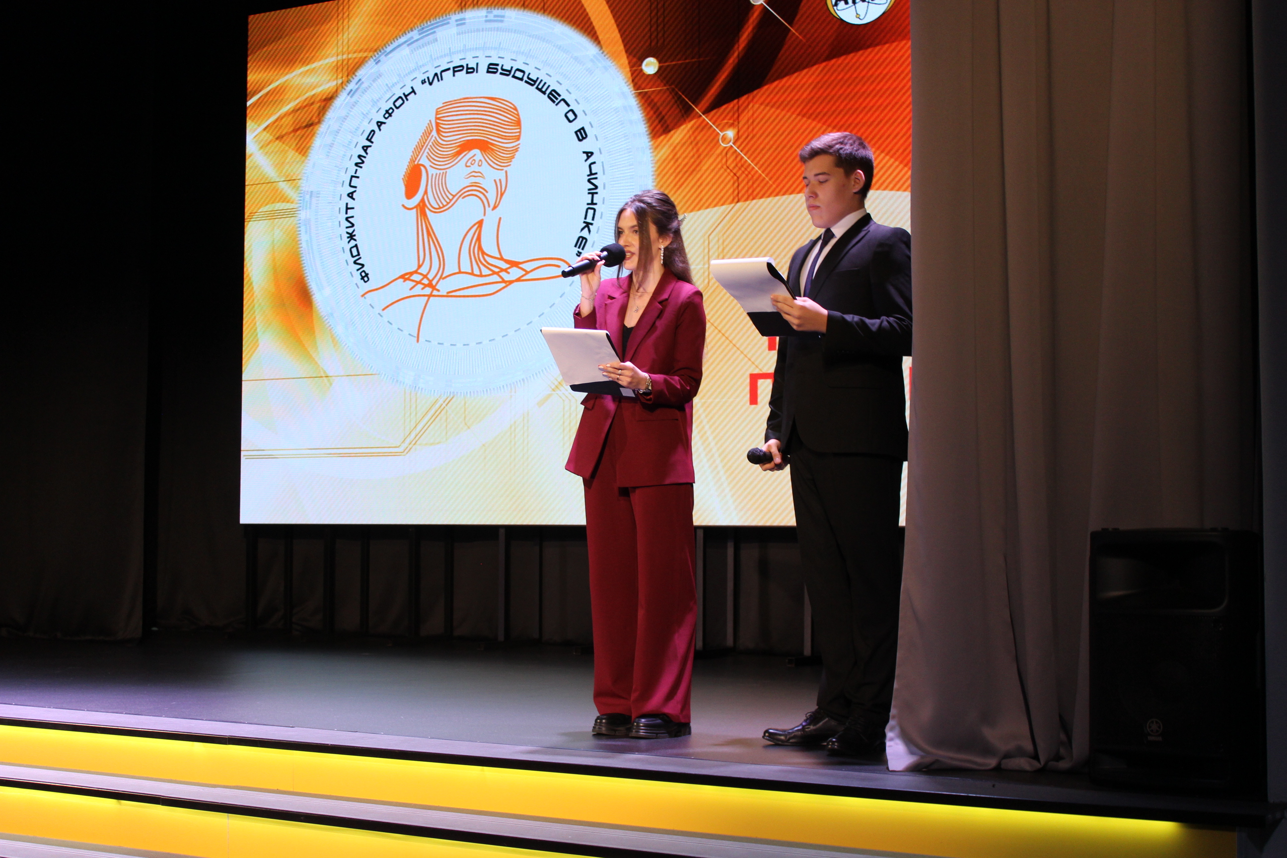 На этом фото два молодых человека находятся на сцене. Один из них – девушка в бордовом костюме, стоит с листом бумаги в руке и произносит речь, а другой – парень в черном костюме и белой рубашке, также с листом в руке, слушает. На заднем плане большая проекция с изображением человека с символами, окруженная яркими цветами, создающими атмосферу торжественного мероприятия.