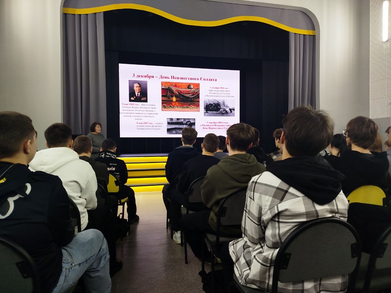 На первом изображении представлена сцена, на которой проходит мероприятие. В центре внимания — человек, стоящий на сцене и обращающийся к аудитории. Сзади него большой экран, на котором отображаются слайды с текстом и изображениями. На экране можно увидеть фотографии и описание. Вся сцена оформлена в светлых тонах, стены серые, а вдоль верхней части экрана проходит жёлтая полоса. Аудитория состоит из людей, сидящих спиной к камере, в основном в тёмной одежде.